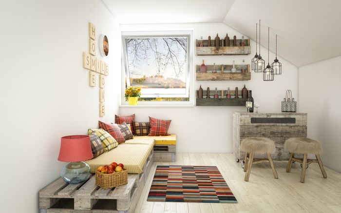 A white room overlooks an autumn hillside scene. A futon bed is made of repurposed wooden crates topped with yellow cushions and red and brown tartan pillows. Two wooden stools with fur seats sit near the window, beside a dresser made of crates.